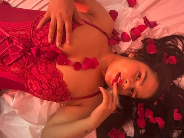Amaia Saenz è sdraiata su un letto cosparso di petali di rosa rossa, indossando un corpetto di pizzo rosso. Le sue mani toccano il petto, le labbra leggermente dischiuse, la testa inclinata. Illuminazione di un rosso acceso, posa seducente.