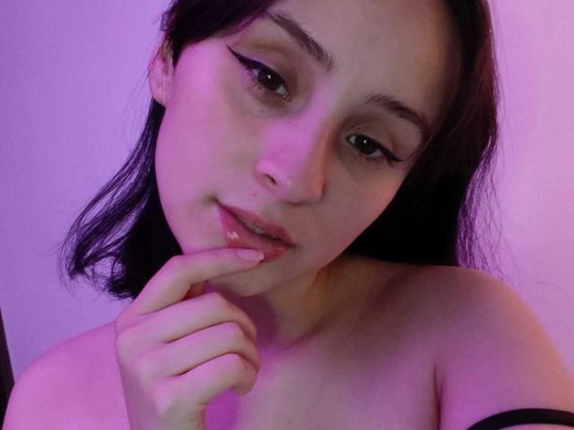 Gros plan sur AnnieLamberth, à la peau claire, avec des cheveux foncés, mordant sa doigt de manière séduisante. L'éclairage violet et rose met en valeur son visage. Épaules nues, décolleté subtil, regard séduisant, lèvres légèrement entrouvertes..