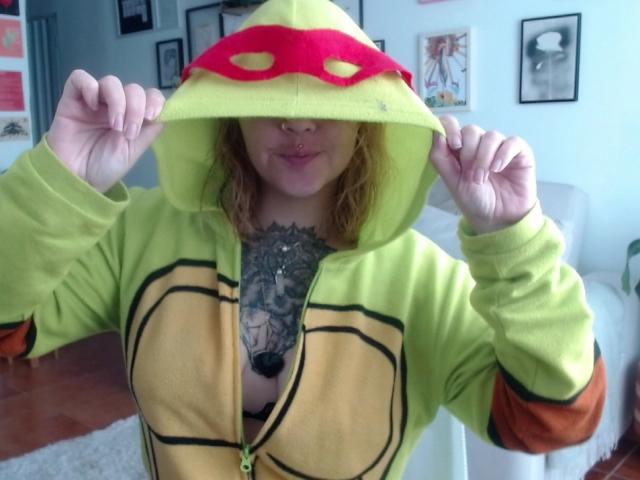 AlinaMatei veste um moletom amarelo com um capuz vermelho em formato de cogumelo, revelando seus seios grandes e uma tatuagem elaborada no peito. As mãos dela seguram a capuz. Quarto claro, com quadros na parede ao fundo..