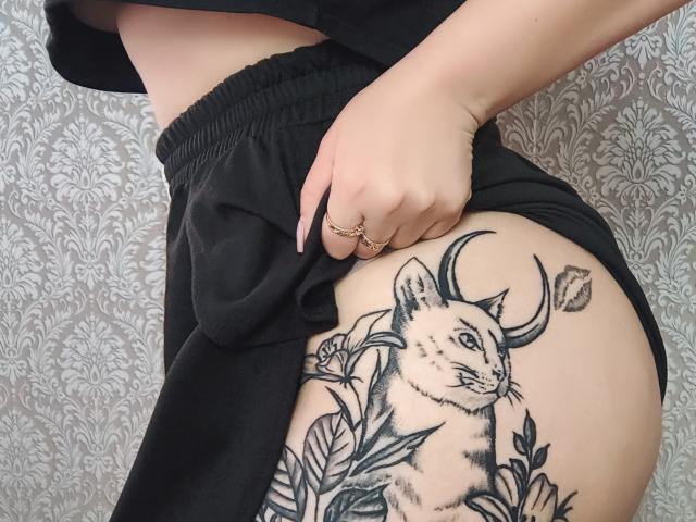 Gros plan du bas du dos et des fesses de Niferttiti, montrant un tatouage noir et blanc détaillé d'un cerf avec des bois. des lianes feuillues sur leur hanche droite. Portant un pantalon noir, la main posée sur la hanche.