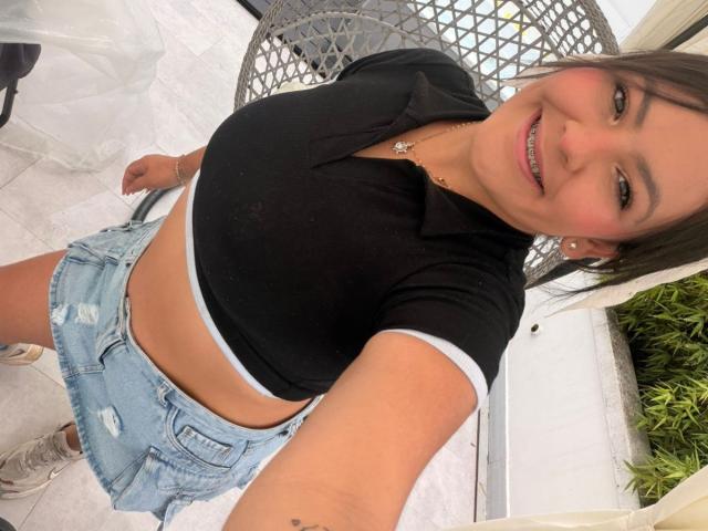 Scarlett Oxford steht leicht geneigt und lächelt in die Kamera. Sie trägt ein schwarzes Crop-Top mit weißer Biese, hellblaue, abgenutzte Shorts und weiße Sneakers. Ihre Hand ruht auf ihrer Hüfte..