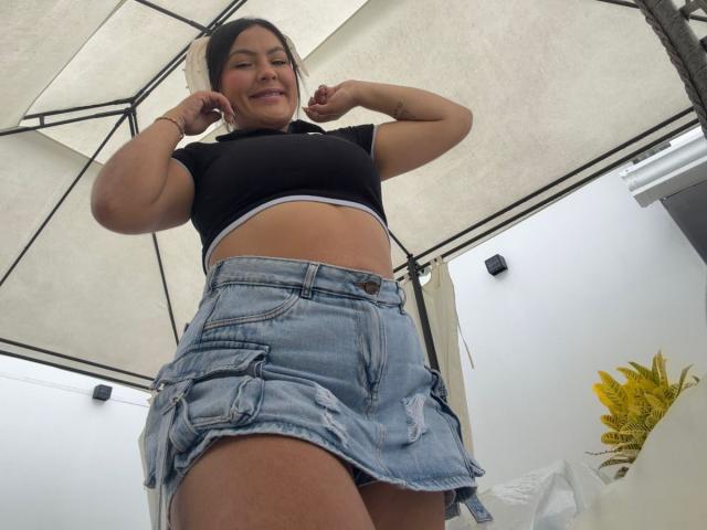 Scarlett Oxford se mantiene erguida con confianza, mirando hacia abajo a la cámara. Ella lleva una camiseta corta negra y una falda vaquera deshilachada, mostrando su abdomen. Ella tiene una sonrisa burlona, con las manos en la cabeza. Plano desde un ángulo bajo..
