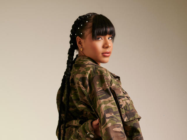 Photographie de KinkyCatalina avec des cheveux tressés foncés, portant une veste camouflage, regardant par-dessus son épaule. Son expression est confiante, les yeux légèrement baissés. Fond clair, ombres douces. Extrêmement sexy..