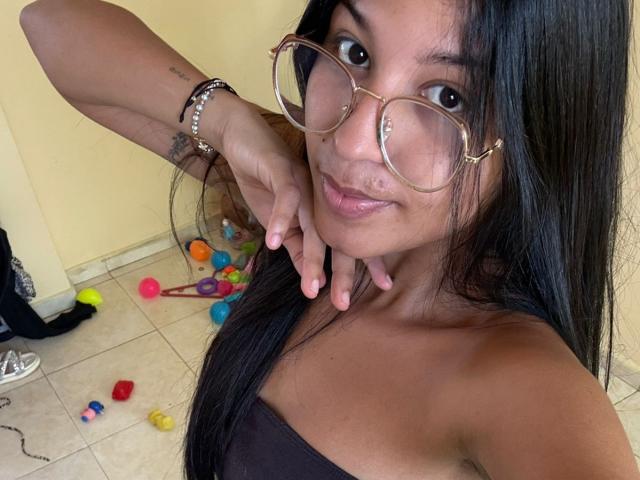 Foto em close-up de BrianaPink, uma mulher atraente com cabelos longos e pretos, usando óculos grandes com armação dourada e um vestido preto. Blusa sem alças. Ela está inclinada para frente, com os dedos perto do queixo, e há brinquedos coloridos no chão..