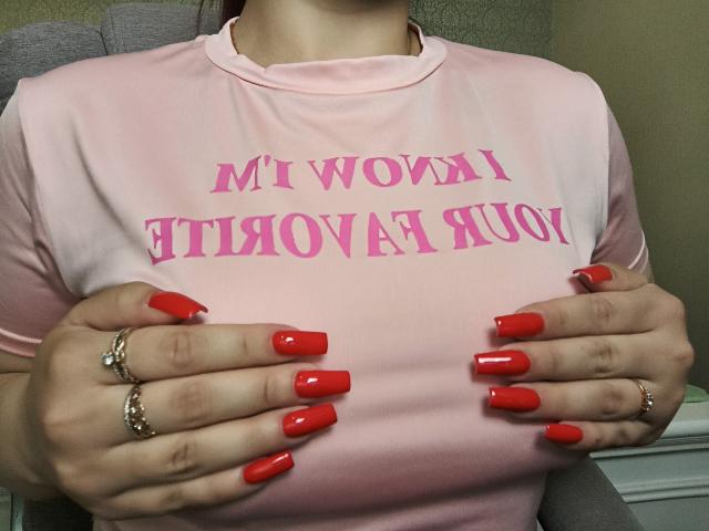 Niferttitis Hände, mit roten, langen Acrylnägeln und mehreren Goldringen geschmückt, umklammern ihr pinkes T-Shirt, auf dem "MOW A ROW A" steht. REIHE A, REIHE. Ihre großen Brüste werden betont. Verdammt sexy..