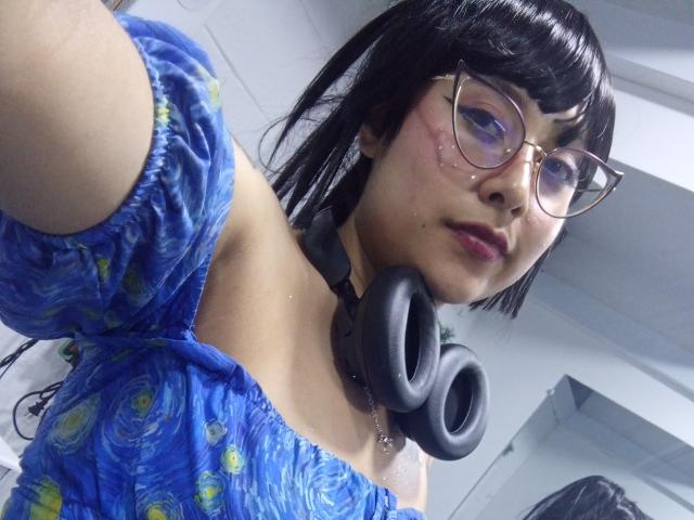 Primer plano de AntonellaHanso con cabello negro, gafas y un vestido azul con estampados amarillos, usando auriculares negros. Alrededor de su cuello, mirando hacia la cámara con una ligera sonrisa. Iluminación interior brillante.