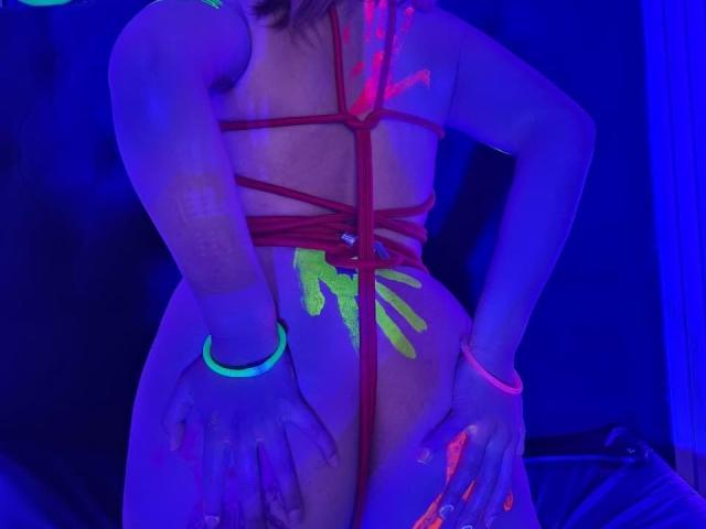 AntonellaHanso, in piedi in una stanza scarsamente illuminata, indossa un'imbracatura di corda rossa, con vernice verde neon e rosa sul corpo. braccialetti al neon luminosi sui polsi, la luce blu che ne evidenzia le curve.