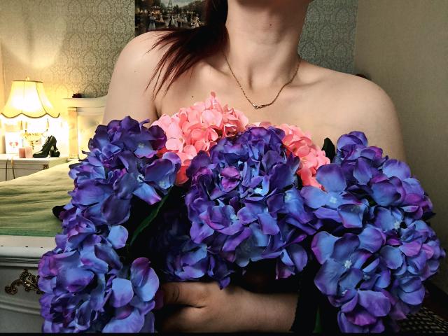 Niferttiti's shoulders and upper chest are covered in a bunch of vibrant purple and pink hydrangeas. Her neck and collarbone are visible, with a warm lamp in the background.