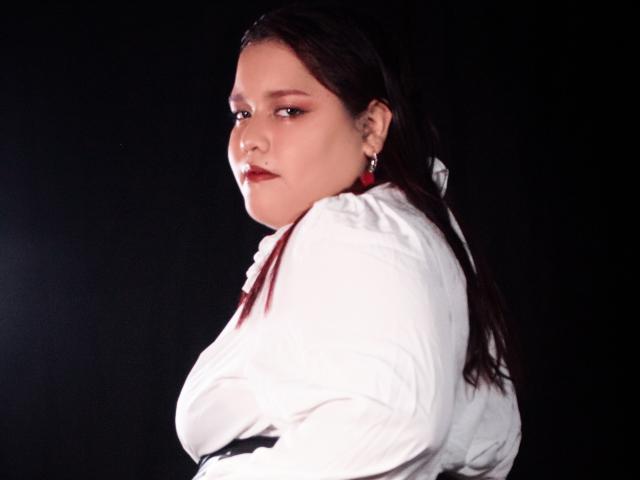 Fotografia de AleSweet, uma mulher curvilínea com cabelos longos e escuros, vestindo uma camisa branca, olhando por cima do ombro. Expressão sensual. Alto contraste, fundo escuro, tensão sexual.