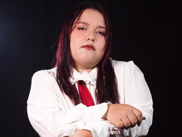 Fotografia de uma jovem, AleSweet, com cabelos longos e pretos, vestindo uma camisa branca e gravata vermelha, braços cruzados, expressão intensa. expressão, fundo preto, contraste marcante, alta saturação. Olhar muito sério..