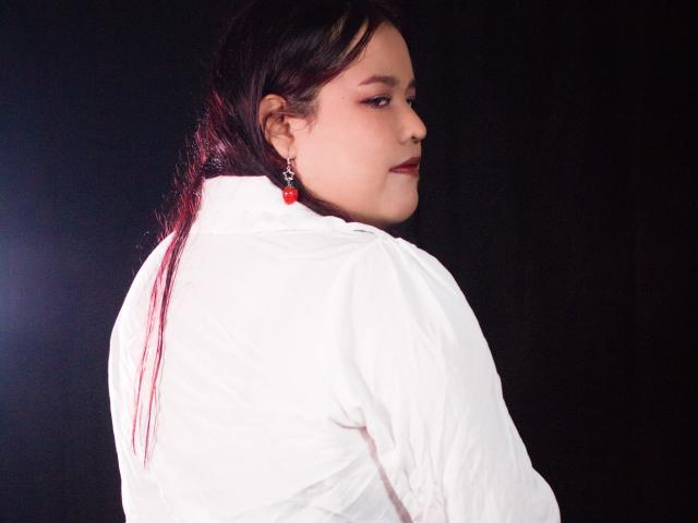 Photograph of AleSweet with long dark hair, white shirt, red earrings, looking back over shoulder, dark background, stark contrast, shadowed face, slight smile, intense gaze, sensual, confident pose.