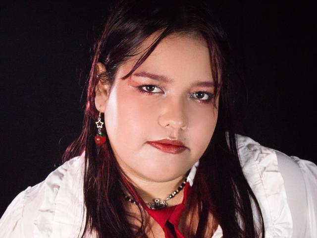 Fotografía de primer plano de AleSweet, una niña regordeta con cabello largo y negro, que lleva una camisa blanca y una corbata roja, y aretes negros con gemas rojas, expresión intensa, fondo oscuro, ligero efecto de brillo..