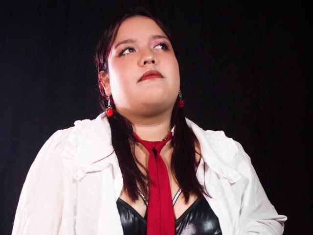 Photograph of AleSweet with light skin, dark hair, and red earrings, wearing a white jacket over a black leather top, red tie, and looking upward with a slightly flushed face. Fucking intense gaze.