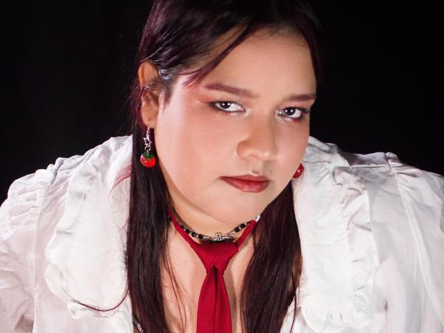 Close-up foto van AleSweet met bleke huid, donker steil haar, rode lippenstift en rode oorbellen. Ze draagt een witte blouse met een rode sjaal, heeft een intense blik en een lichte grijns. Achtergrond zwart.