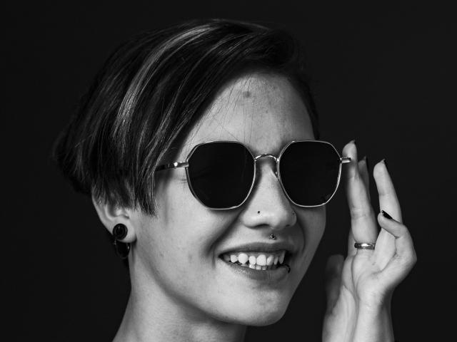 Photo en noir et blanc de LissyLuxe, avec des cheveux courts et foncés, des lunettes de soleil rondes, souriante, une main près du visage, des bouchons d'oreille noirs, et un nez. piercing, bague au doigt, contraste saisissant, sexy, ambiance confiante.