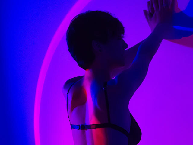 Silhouetted profile of LissyLuxe, facing right, arms raised, wearing a black bra, illuminated by vibrant blue and purple neon light. Shadows highlight curves, creating a provocative, sensual atmosphere.