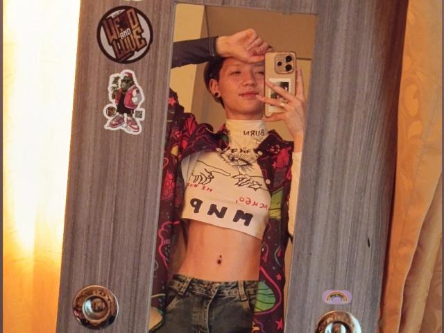Autoportrait de LissyLuxe, avec une coupe de cheveux courte, tenant un téléphone, portant une chemise rouge à carreaux ouverte et un crop top blanc avec l'inscription "PUNK". texte, ventre apparent, jean bleu, autocollants sur le miroir, tension sexuelle, pose décontractée.