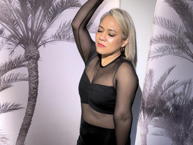 CamilaNoer with blonde hair, closed eyes, and a slight smile, wearing a black sheer top and black skirt, hand raised, palm tree pattern in the background, sexual tension.