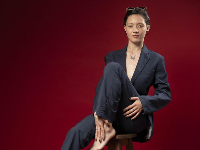 LissyLuxe sits on a stool against a deep red background, wearing a black suit with a V-neck, legs crossed, one hand on her knee, the other on the stool. Fucking sexy.