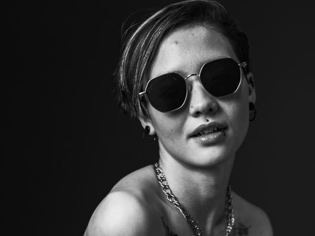 Black-and-white photograph of LissyLuxe with short hair, wearing round sunglasses, a chain necklace, and black ear gauges. LissyLuxe's expression is slightly seductive, with a hint of a smirk. High contrast.