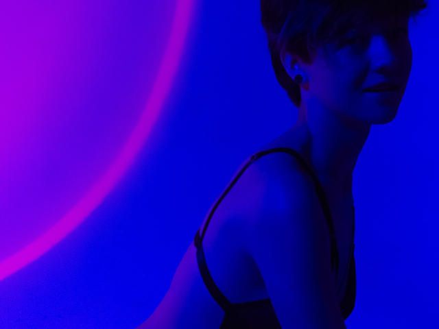El rostro de LissyLuxe está parcialmente oculto, vista de perfil, vistiendo un top negro sin tirantes. La luz de neón azul y púrpura crea una tensión sexual, el cabello corto de LissyLuxe, su mirada intensa, sus facciones en sombra. Composición audaz y provocadora.