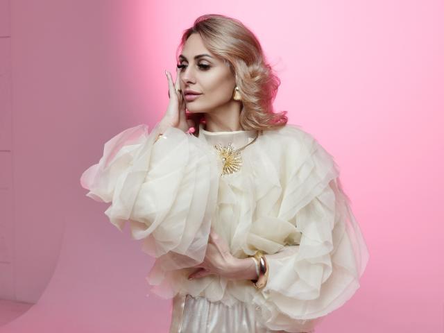 CrystalSky staat afgebeeld tegen een roze achtergrond, en draagt een witte top met ruches en gouden accenten. Ze raakt haar wang aan met één hand, en ziet er verleidelijk uit. Blond haar, zacht licht, zelfverzekerde pose..