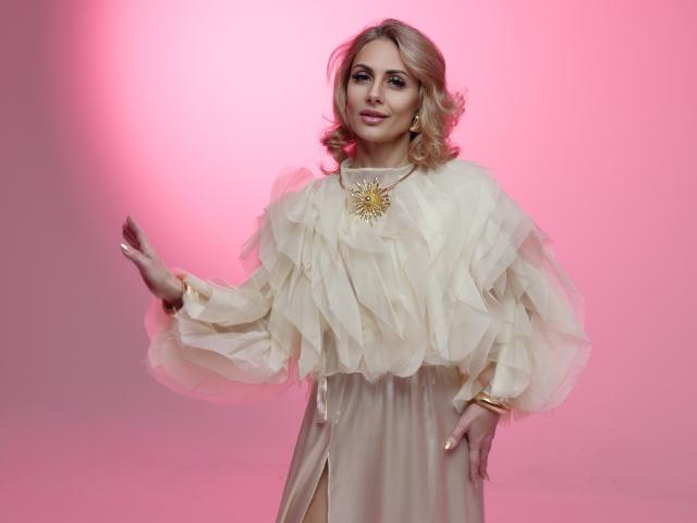 CrystalSky, une femme blonde, se tient devant un fond rose dégradé. Elle porte un haut blanc, à volants et aux manches bouffantes, avec un collier en or, et une main levée. Pose incroyablement séduisante, élégante et pleine d'assurance..