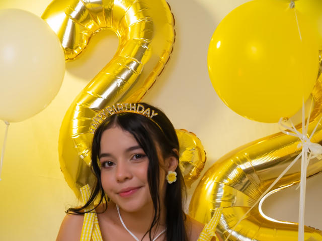 LissyCarter, jeune femme aux longs cheveux noirs, portant des boucles d'oreilles jaunes et un haut jaune, entourée de chiffres "2" dorés brillants. ballons et ballons jaunes. Couleurs vives et jaune, ambiance festive
.