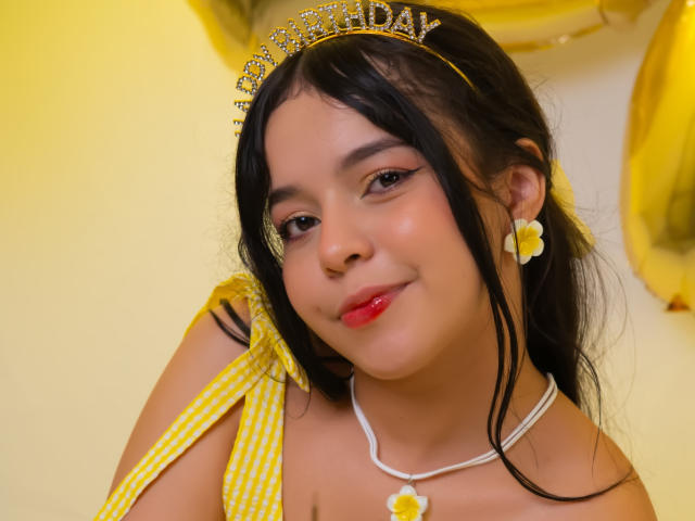 Close-up photo of LissyCarter, young woman with light brown skin, black hair, wearing a glittery yellow tiara, yellow ribbon, yellow earrings, red lipstick, white necklace with yellow pendant, smiling seductively.