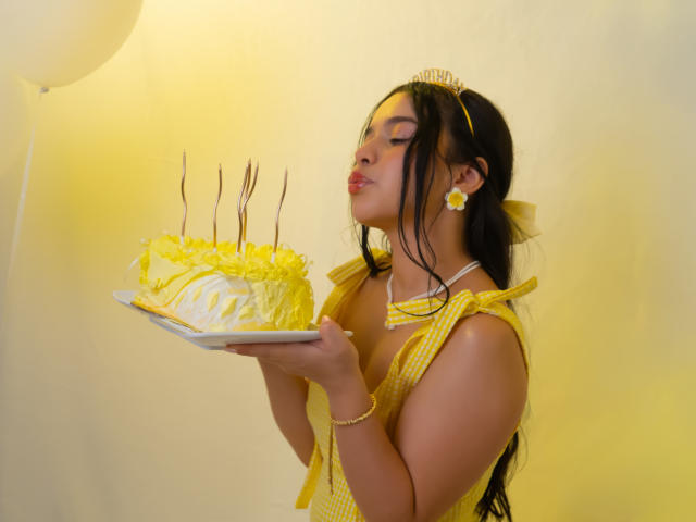 Lissy Carter, con cabello oscuro, vestida con un vestido amarillo y accesorios a juego, sostiene un pastel con glaseado amarillo y velas, ojos cerrados, labios fruncidos, fondo amarillo brillante. Ambiente de cumpleaños genial..