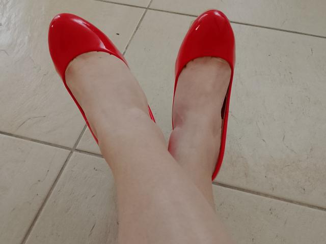 Primo piano delle gambe chiare di Marriam, incrociate alla caviglia, che indossa tacchi alti rossi lucidi su un pavimento piastrellato beige. I tacchi rossi creano un contrasto audace, mettendo in risalto i suoi piedi e le sue gambe..
