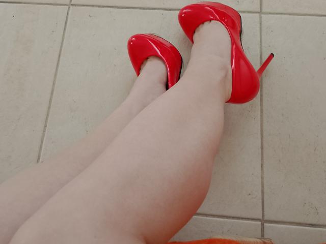 Marriam's pale legs, crossed at the ankles, wear shiny red high heels with stiletto heels on a tiled floor. The bright red shoes create a stark contrast with the pale skin.