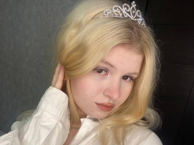 Delphine Hambleton, com cabelo loiro, usando uma tiara prateada, camisa branca e maquiagem discreta, com a mão no cabelo, olhando para a câmera com um leve sorriso. Seus seios não estão visíveis..