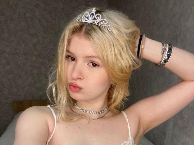 Delphine Hambleton mit blonden Haaren, die eine silberne Tiara, eine Kette und Armbänder trägt, verzog den Mund und hob ihren Arm, wobei sie in einer weißen Spitzenbluse ihre Brüste zeigte..