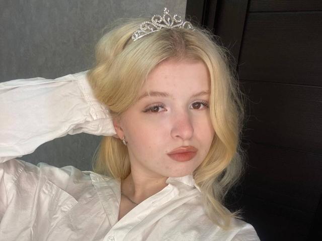 Delphine Hambleton, avec ses cheveux blonds, portant une tiare argentée et une chemise blanche boutonnée, faisant la moue, une main dans ses cheveux, yeux marrons, peau claire, maquillage discret, regardant la caméra..