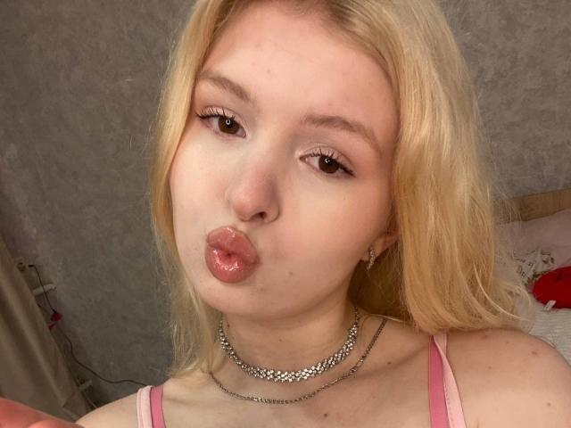 DelphineHambleton with blonde hair, pink top, puffy pink lips, and a silver chain necklace, making a pouty face. She has small breasts and light skin.