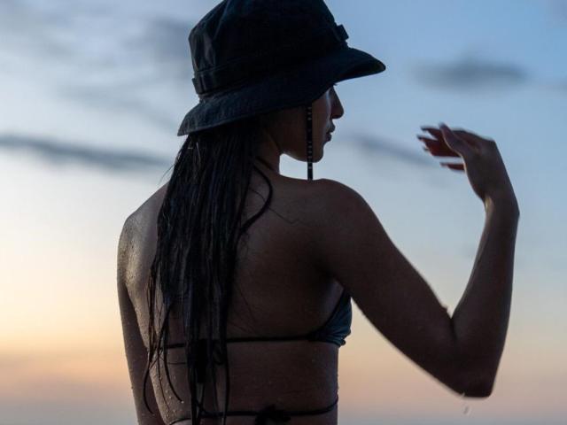 Fotografia in silhouette di MiaaFerrer, vista posteriore, che indossa un cappello bagnato e un top da bikini, lunghi capelli afro bagnati, una mano sollevata. cielo al tramonto con sfumature di blu e arancione, tensione sessuale, fisico slanciato..
