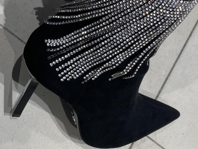 Gros plan du talon aiguille noir de DeesseBleue, orné de perles et de paillettes. Des ombres contrastées sur le sol carrelé. La pointe effilée de la chaussure et son talon aigu créent une silhouette suggestive..