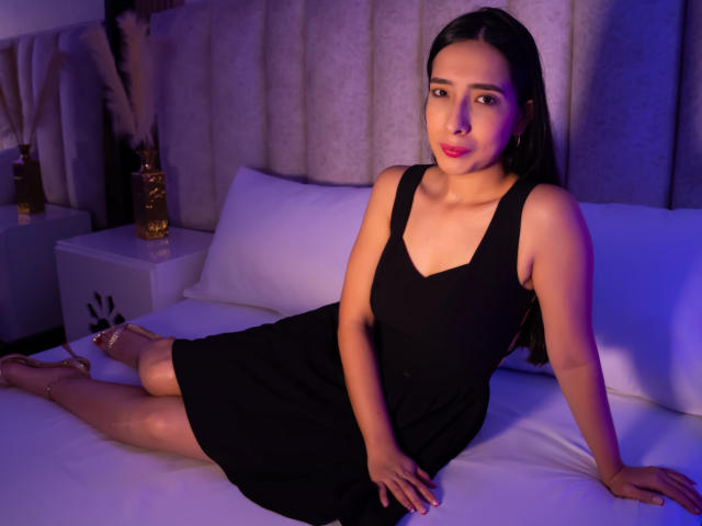 Ivanka Clark, eine attraktive lateinamerikanische Frau mit langen, schwarzen Haaren, sitzt auf einem Bett in einem schwarzen Kleid, die Beine gekreuzt, die Hand auf Weiß. ein Kissen, beleuchtet von blauem und violettem Licht. Ihr roter Lippenstift bildet einen Kontrast zu ihrer hellen Haut..