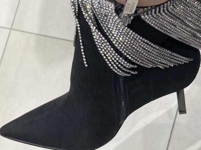 Primer plano de una bota negra de tacón alto con adornos de lentejuelas plateadas en el borde superior. La punta del botín es afilada y está orientada hacia arriba, mostrando una punta de tacón de metal. Suelo de baldosas blancas.