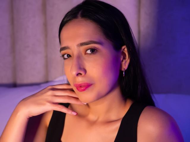 Photograph of IvanaClark, a young woman with long black hair, wearing a black tank top, looking seductively at the camera with her hand touching her chin, glowing pink lips. Purple and blue lighting.