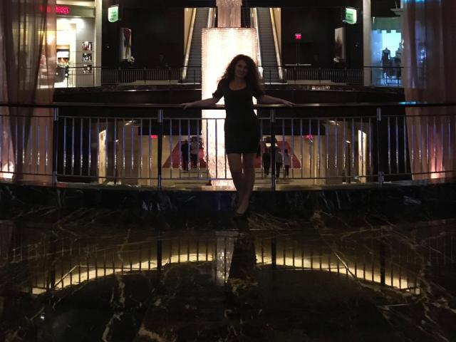 Silhouette of DonJuanundEva-hot standing in front of a brightly lit building at night. She's wearing a short dress, arms outstretched, with wet marbled ground reflecting light. Fucking intense.