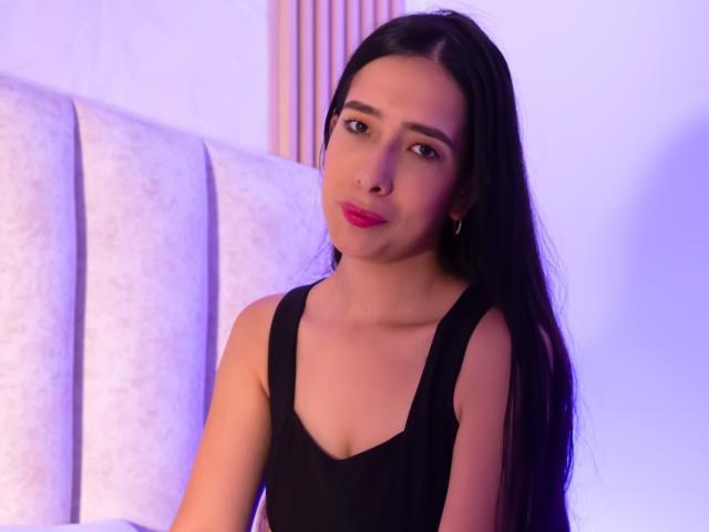 Photograph of IvanaClark, a slim woman with long black hair, wearing a black tank top, sitting against a white cushioned background with blue lighting. Her expression is slightly seductive.