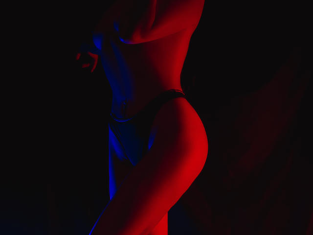 Silhouetted HelenHard, lit by red and blue lights, stands in profile. She's topless, wearing black thong, emphasizing her toned ass and slim waist. Red light highlights her right side.
