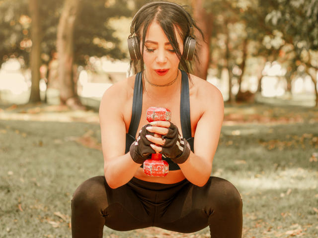 HelenHard squats in a park, holding a red, glittery object between her black-gloved hands. She wears black sports bra and pants, headphones over messy black hair. Focused, sexual tension.