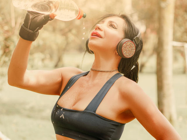 HelenHard drinking from a plastic bottle, sweat on her chest, black sports bra, black gloves, large round headphones, forest background. Sunlight highlights her skin, red lipstick, intense gaze.