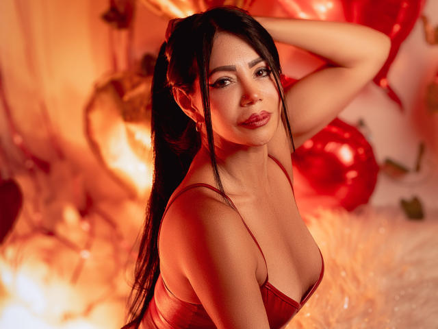HelenHard with long black hair, red lingerie, and red lips, posing seductively with one hand behind her head. Bright red and orange light, red ornaments, and blurred background. Fucking sexy.