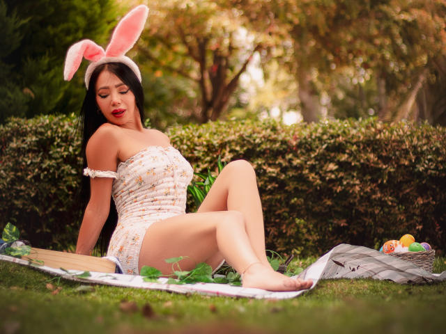 HelenHard sits on grass with legs spread, wearing a white off-shoulder top and pink bunny ears, eyes closed, lips red. Basket with eggs and blanket nearby. Sunny outdoor setting.