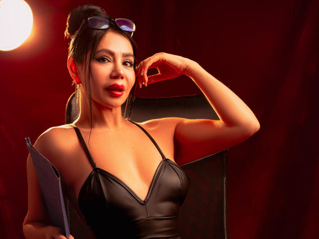 HelenHard with dark hair in an updo, wearing a black leather dress, red lipstick, and black sunglasses on her head. Her right arm raised, hand touching her head. Red lighting.