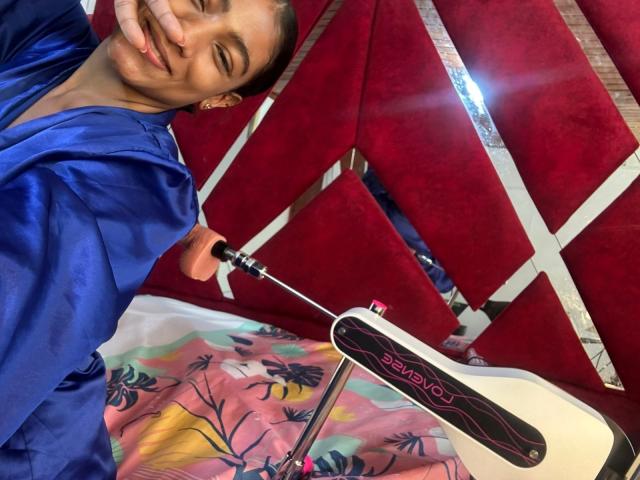 NatalyBlonder with closed eyes, smiling, wearing a blue shirt, holding a black and white "Super Eight" camera, red fabric background, colorful patterned cloth below, casual, relaxed pose.